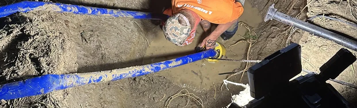 A Raymond plumber in an orange shirt working on blue water line in a flooded trench